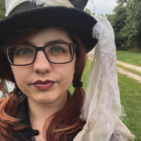 Photograph of Melody in front of a grassy dirt road, wearing a beat-up top hat with tattered wrap hanging down to shoulder. She has low pigtails, glasses, and dark lipstick.